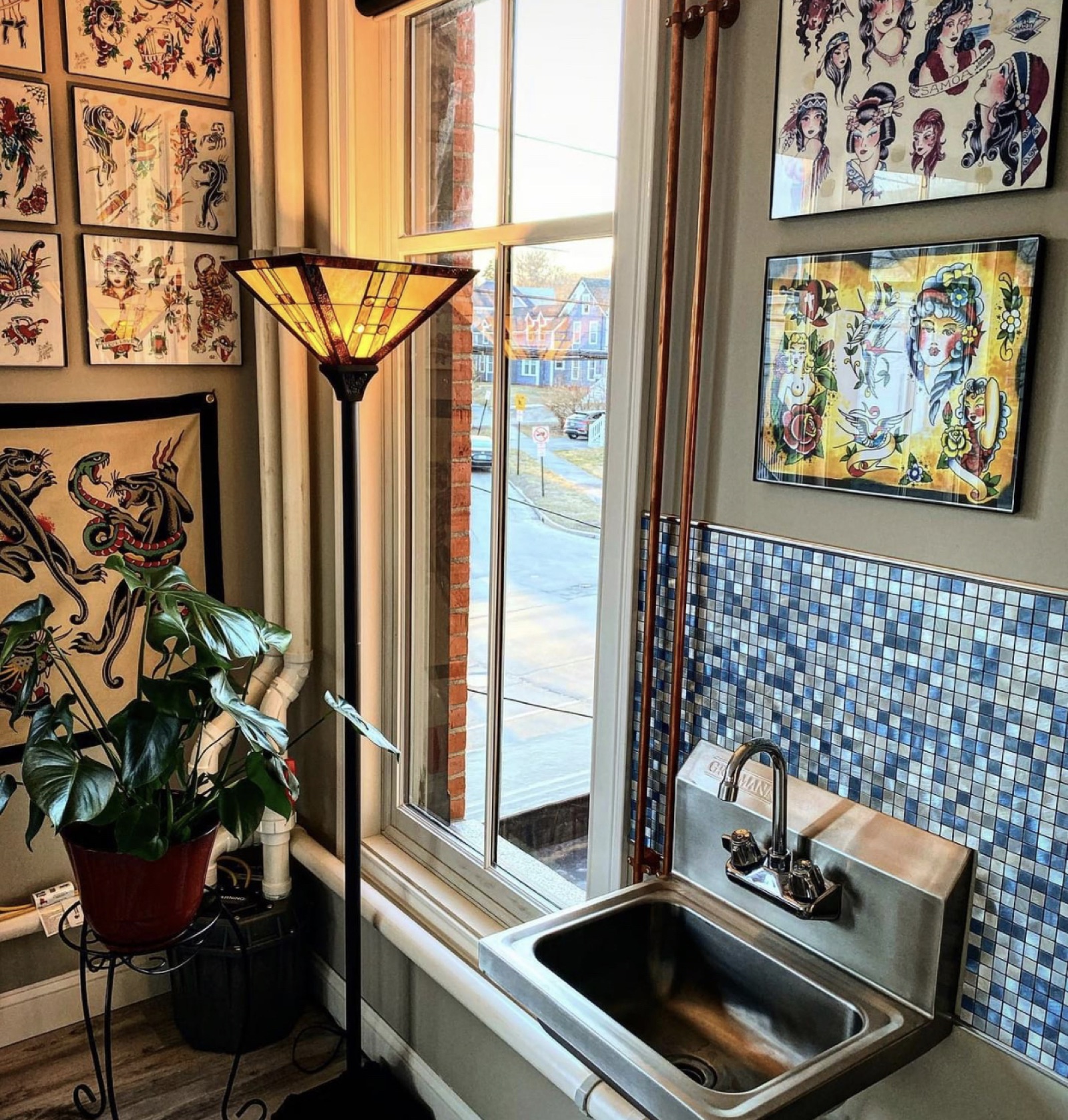 Studio corner with framed traditional flash art, a stained-glass lamp and a plant by the window