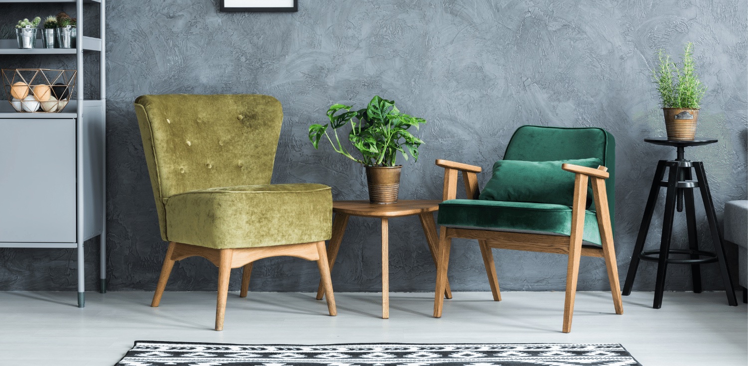 A green velvet wingback chair and a green-upholstered mid-century lounge chair around a round walnut side table in a softly lit room